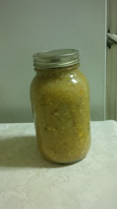 A mason jar on a white counter against a white background.  It is full of thick pale tan liquid with pieces of corn and green flecks suspended in it.